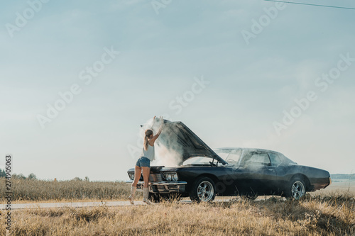 Young female standing near overheated car in the field, bright sunlight, steam under the hood