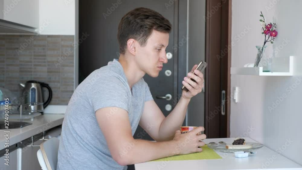 Portrait of young man is having a breakfast. He is drinking cup of tea and browsing phone sitting in a small kitchen. Portrait of young man is having a breakfast. He is drinking cup of tea and browsing phone sitting in a small kitchen.