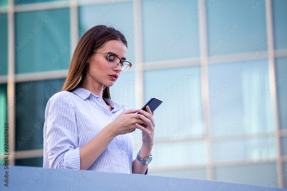 Executive business woman looking at mobile smartphone in the street with office buildings in the background Executive business woman looking at mobile smartphone in the street with office buildings in the background