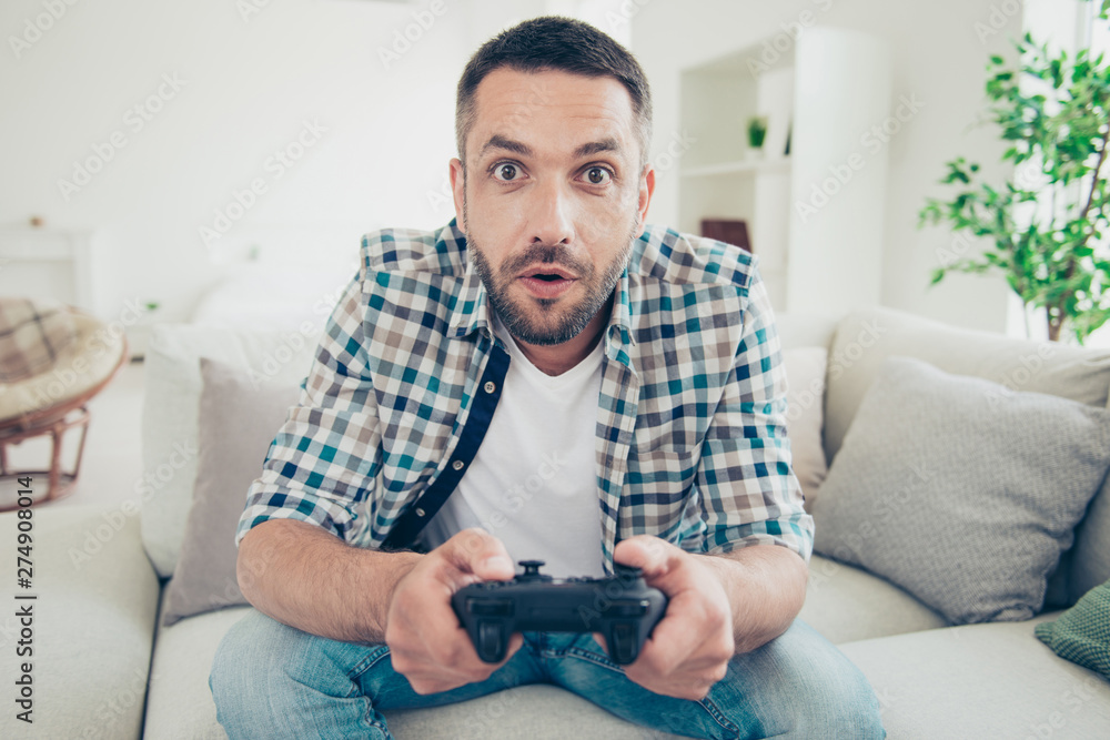 Close up photo of focused handsome mature guy hold hand have play station game pad dressed plaid denim jeans sit divan bearded amazed big apartment Close up photo of focused handsome mature guy hold hand have play station game pad dressed plaid denim jeans sit divan bearded amazed big apartment