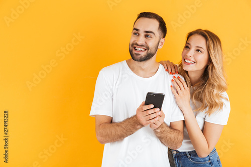 Image of satisfied couple man and woman smiling and using cellphone while looking aside at copyspace together