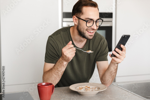 Happy young handsome man posing at the kitchen at home have a breakfast using mobile phone.