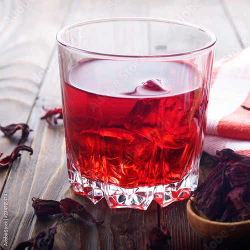 Closeup view at glass of tea with ice and spoon of dry hibiscus petals on wooden table background