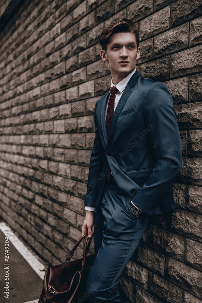 Handsome businessman with bag in his hand. Confident young man in full suit looking away while standing outdoors with cityscape in the background Handsome businessman with bag in his hand. Confident young man in full suit looking away while standing outdoors with cityscape in the background