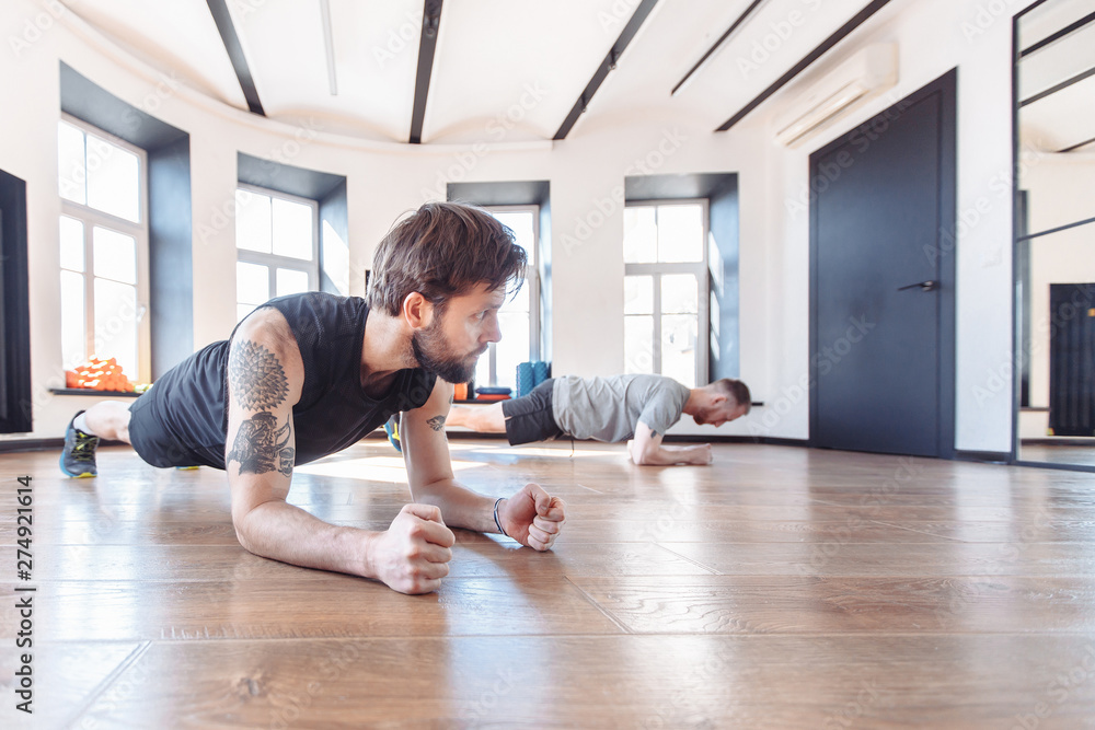 Two friends men in good physical shape doing workout together in the gym. Concept of team spirit and joint visits to the hall for support. Side view of two young sportman in sportswear doing plank Two friends men in good physical shape doing workout together in the gym. Concept of team spirit and joint visits to the hall for support. Side view of two young sportman in sportswear doing plank