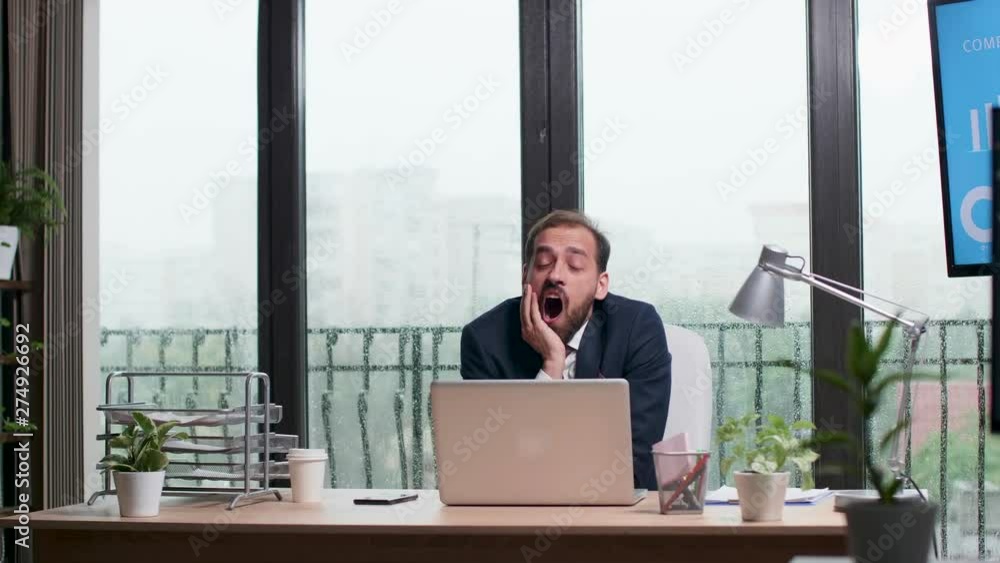 Tired businessman alone in the office yawns while a heavy rain is going outside Tired businessman alone in the office yawns while a heavy rain is going outside