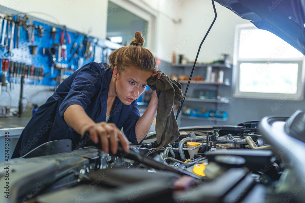 Making some adjustments to her car. Portrait Of Female Auto Mechanic Working. Mechanic repairs the engine of a car in her workshop. Mechanic examining under hood of car at the repair garage Making some adjustments to her car. Portrait Of Female Auto Mechanic Working. Mechanic repairs the engine of a car in her workshop. Mechanic examining under hood of car at the repair garage