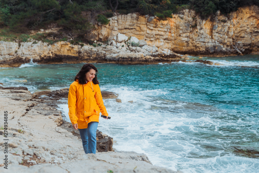 woman in yellow raincoat walking by rocky beach looking at see