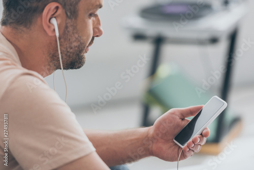 adult man listening music in earphones while holding smartphone with blank screen