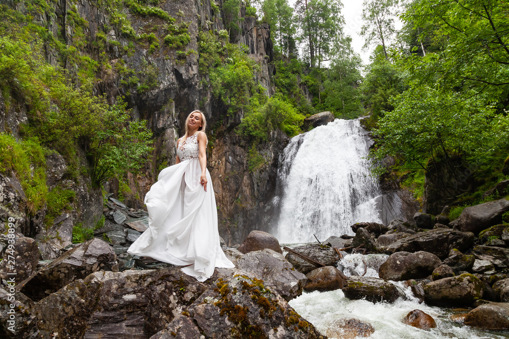 A young blonde girl in an elegant pose hold in arms hem of a boudoir dress in the mountains against a waterfall and stones smiling like a ballerina on a summer day in Altai green trees
