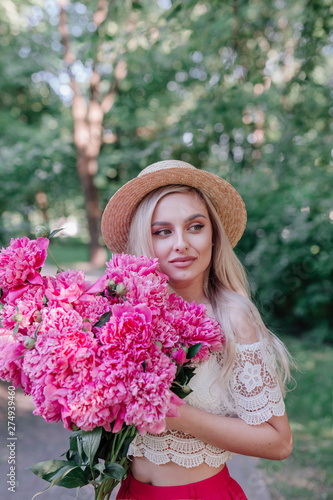 Beautiful young woman with long blond hair posing with bouquet of peonies in a garden