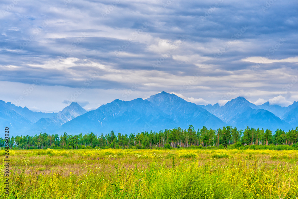 Picturesque summer landscape of mountain range with peaks, valley with green grass, grove and cloudy sky. Natural background with space for text. Eastern Sayan, Tunka National Park, Buryatia, Russia