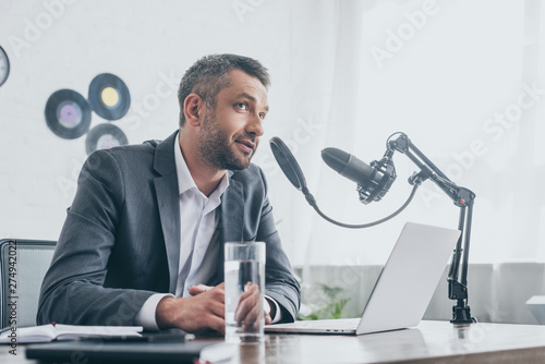 smiling radio host speaking in microphone while sitting near laptop in broadcasting studio