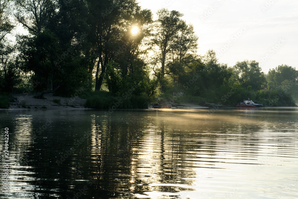 Dawn. The sun shines through the trees and the sunlight reflects in the river. On the river there is a fog. There are boats near the shore. Concept - travel. Dawn. The sun shines through the trees and the sunlight reflects in the river. On the river there is a fog. There are boats near the shore. Concept - travel.