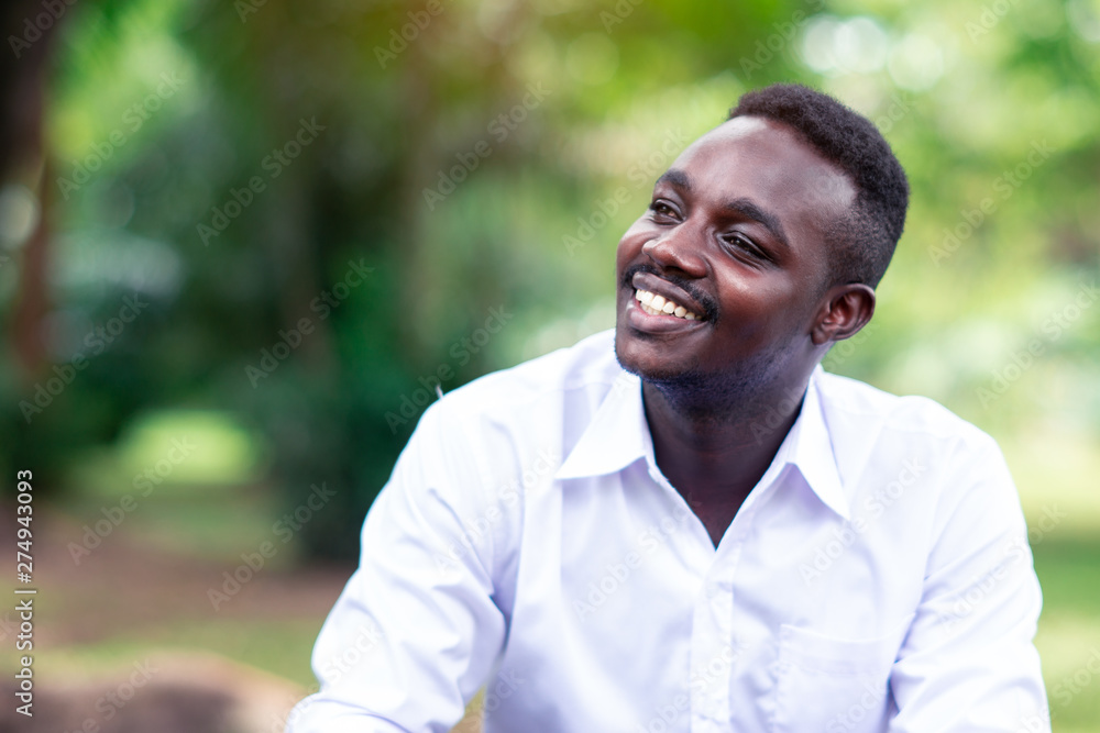 African business man in white shirt smiling and sitting outside with green trees on background. African business man in white shirt smiling and sitting outside with green trees on background.