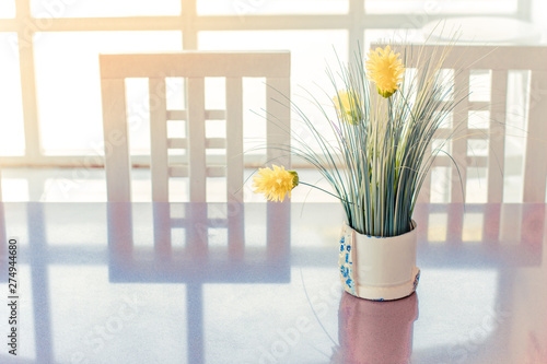 light minimalism interior wallpaper picture with yellow flowers on a smooth kitchen table surface with reflection from white chairs in sunny light through window, copy space 