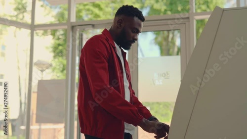 Side view of young stylish Afro-American man inserting credit card in ATM machine, entering code, withdraws cash, machine is broken, money fly out. Shock, problem, stress