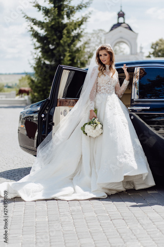 Elegant Bride with Flower Bouquet near Limousine. Sharming Cute Young Woman Dressed in White Wedding Clothing and Jewelry Diadem. Girl Standing near Black Bridal Car on Nature Background