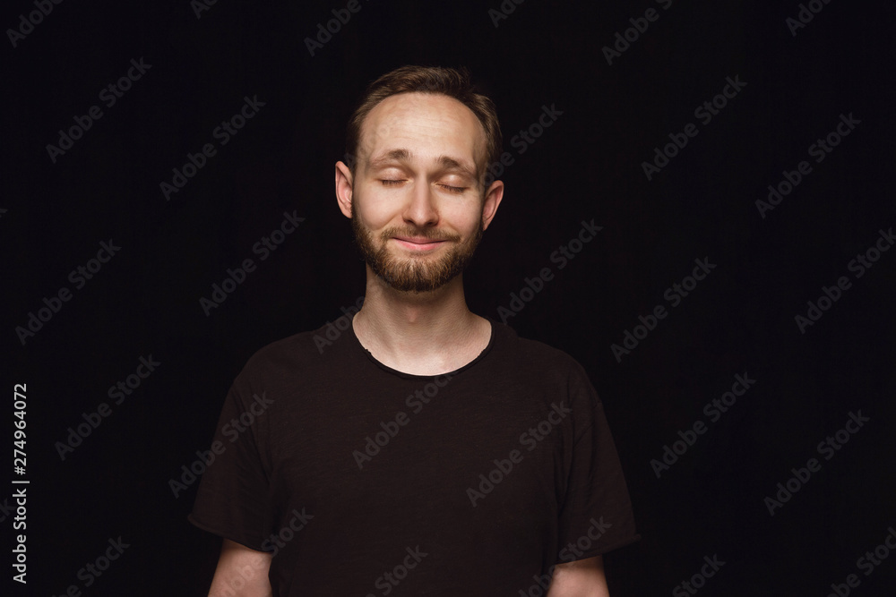 Close up portrait of young man isolated on black studio background. Photoshot of real emotions of male model with closed eyes. Thinking and smiling. Facial expression, human emotions concept.