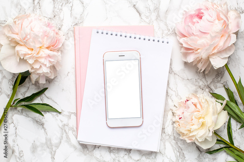 Mobile phone with a white and pink notebook and piony flowers on a marble background