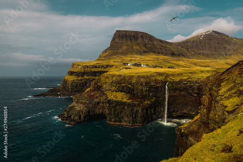 View onto famous Gasadalur waterfall from viewpoint during a sunny spring day with snow-covered mountain peaks and dark blue sea (Faroe Islands, Denmark, Europe)