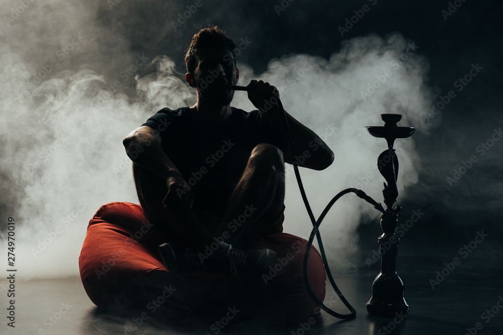 silhouette of man sitting on bean bag chair and smoking hookah in darkness silhouette of man sitting on bean bag chair and smoking hookah in darkness