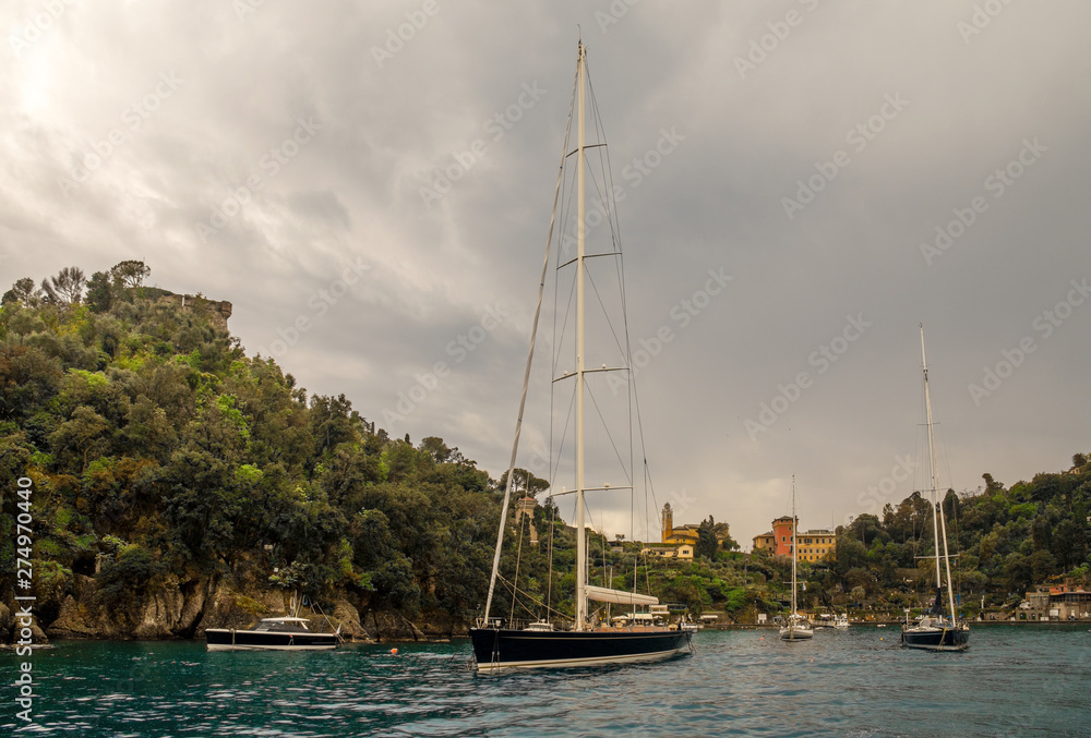 Obraz premium Malowniczy widok z morza na zatokę Portofino, słynną wioskę rybacką na Riwierze Liguryjskiej, z luksusowymi żaglówkami i jachtami zakotwiczonymi w małym porcie w pochmurny dzień, Genua, Liguria, Włochy