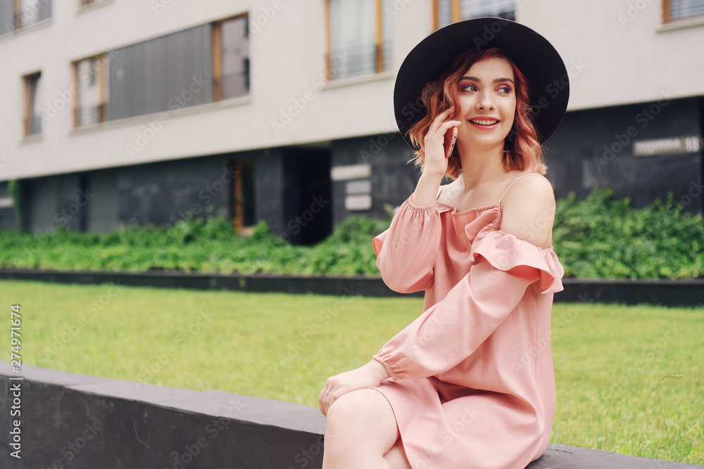 Beautiful girl in a pink dress sitting near high-rise buildings . Girl speaks by phone. Woman with pink hair in a black hat with phone. The girl in the elite area sits on the grass.Girl travels roamin