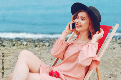 Beautiful girl in a hat with pink hair and beautiful makeup lying on a lounger against the beach and the ocean.Woman is talking on the phone on the beach, freelancing, roaming, holidays.