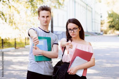 Two angry and bewildered friends holding books, notes and other learning materials gesturing thumbs down in the street near old conventional university