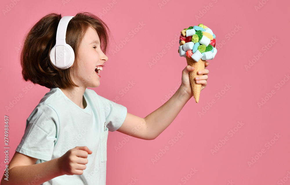 Happy teen girl kid walks in headphones sings with a big ice cream in waffles cone with tasty toppings give it on pink Happy teen girl kid walks in headphones sings with a big ice cream in waffles cone with tasty toppings give it on pink