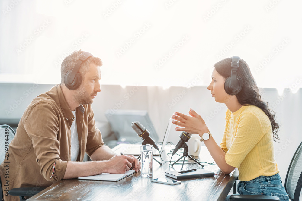 Obraz premium two serious radio hosts talking while sitting near microphones in broadcasting studio