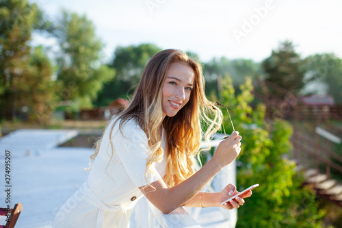 Fototapeta Naklejka Na Ścianę i Meble -  Elegant charming woman texting romantic message on mobile phone. Portrait of luxary lady in glasses enjoying summer sunset in cafe outdoor. Communicating by smartphone in travel..