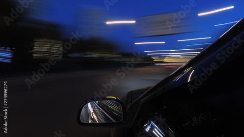 The dark-colored car is moving fast on the illuminated road of the night city, on a blurry flying background one can see a part of the car with a mirror