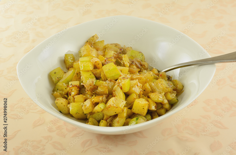 Yellow summer squash and zucchini with onion side dish in white serving bowl with serving spoon on tablecloth Yellow summer squash and zucchini with onion side dish in white serving bowl with serving spoon on tablecloth