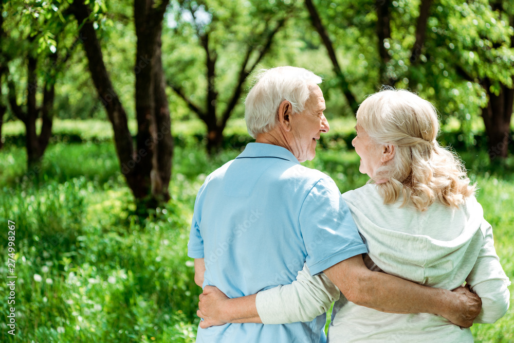 smiling pensioner hugging happy senior wife while standing in green park smiling pensioner hugging happy senior wife while standing in green park