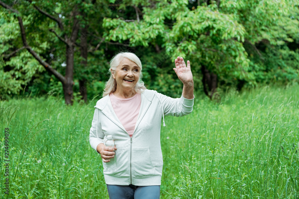 cheerful senior woman with grey hair waving hand and holding bottle with water