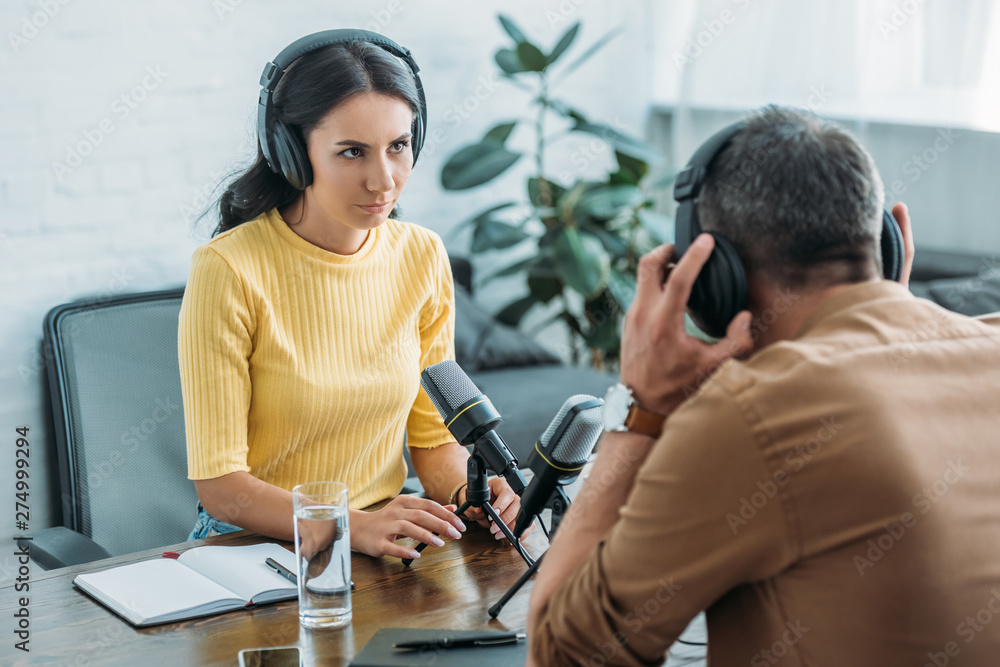 Fototapeta premium attractive serious radio host looking at colleague while sitting at workplace in studio