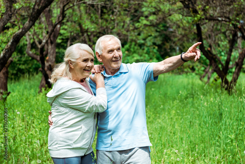 cheerful retired man pointing with finger near happy wife with grey hair