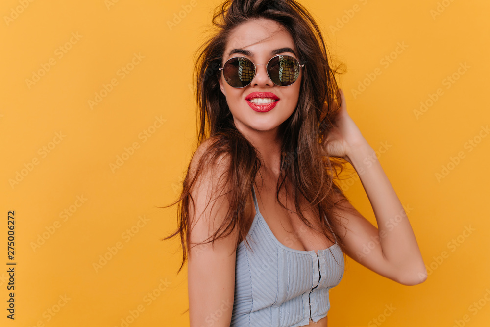 Pleased young woman with inspired smile playing with dark hair. Indoor portrait of stunning tanned girl in black sunglasses isolated on bright background. Pleased young woman with inspired smile playing with dark hair. Indoor portrait of stunning tanned girl in black sunglasses isolated on bright background.