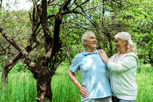 cheerful senior man standing with hand on hip near wife and green tree