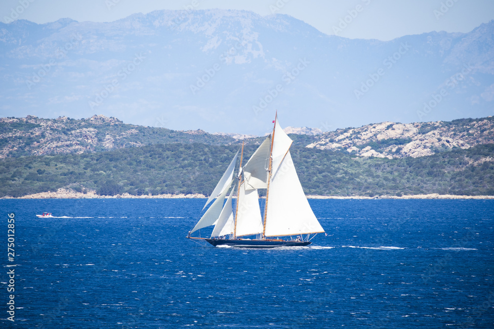 Naklejka premium Stunning view of a beautiful sailboat sailing on the Mediterranean sea that bathes the coasts of Sardinia. Maddalena Archipelago National Park in the background, Sardinia, Italy.