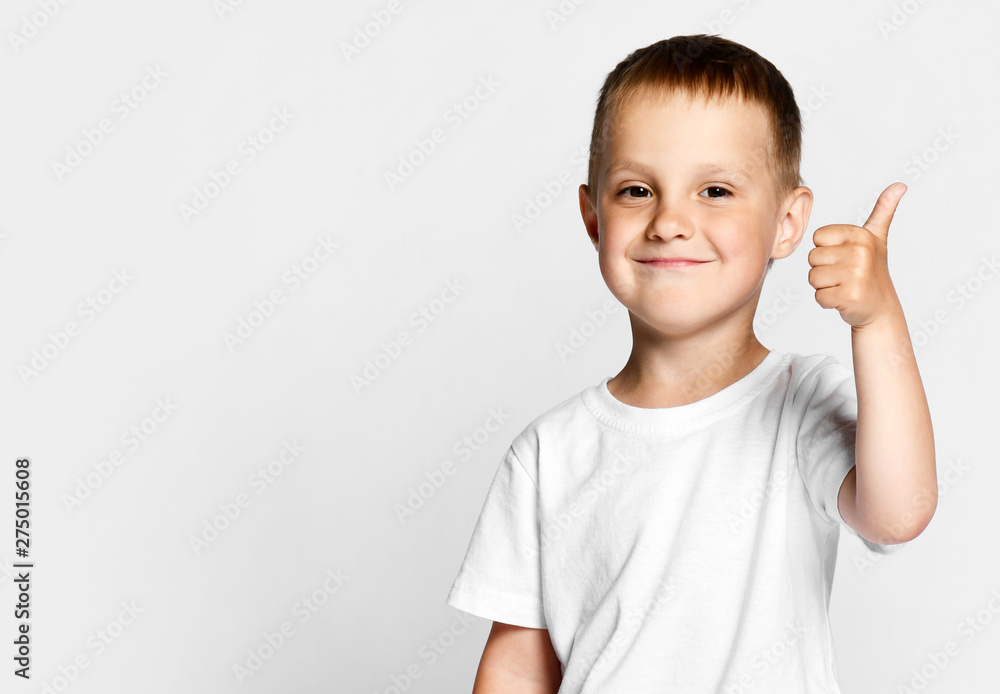 Little cute boy preschooler 4 years old in a white T-shirt and gray pants on a light background, happily looking at the camera, showing a thumb sign cool Little cute boy preschooler 4 years old in a white T-shirt and gray pants on a light background, happily looking at the camera, showing a thumb sign cool
