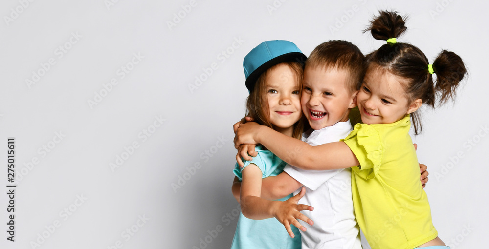 Studio portrait of children on a light background: full body shot of three children in bright clothes, two girls and one boy. Triplets, brother and sisters. hugging on camera. Family ties, friendship Studio portrait of children on a light background: full body shot of three children in bright clothes, two girls and one boy. Triplets, brother and sisters. hugging on camera. Family ties, friendship
