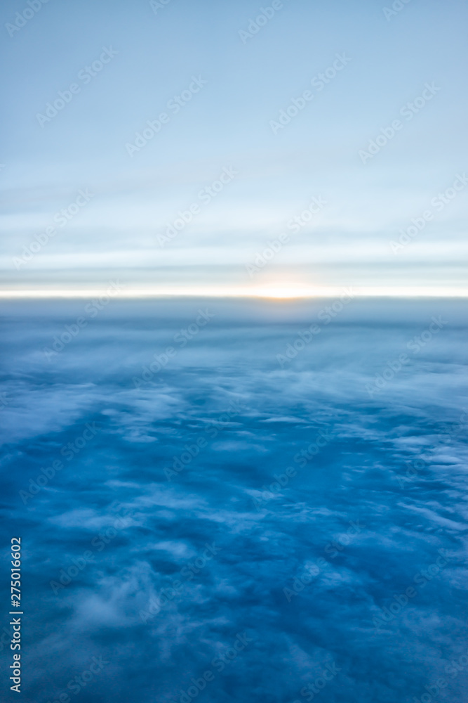 Blue sky with view from airplane window high angle from plane and sun setting above ocean horizon blurry background