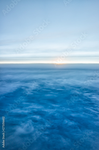 Blue sky with view from airplane window high angle from plane and sun setting above ocean horizon blurry background