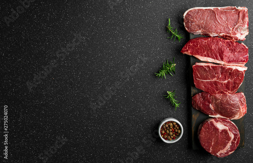 Selection of raw beef meat food steaks against black stone background. New york striploin steak, top blade, rib eye, and other cuts of meat.