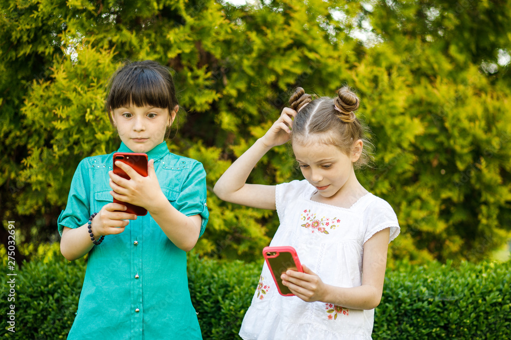 Two cute girls of school age photograph themselves on the phone near the green trees. Children are having fun Two cute girls of school age photograph themselves on the phone near the green trees. Children are having fun