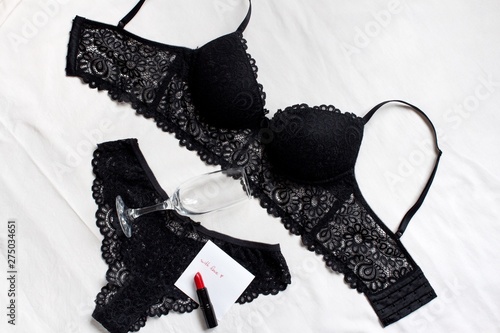 black set of women's underwear, a champagne glass, red lipstick and love note on a bed with white sheet