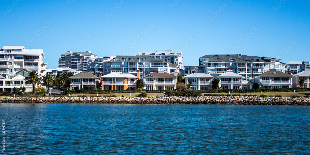 Harbourside condominium apartments behind free standing houses with grass frontage, stone retaining wall and blue river against blue sky